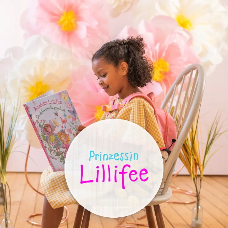 Ein Mädchen sitzt auf einem Stuhl vor einem Hintergrund mit Papierblumen und liest in einem Buch von Prinzessin Lillifee