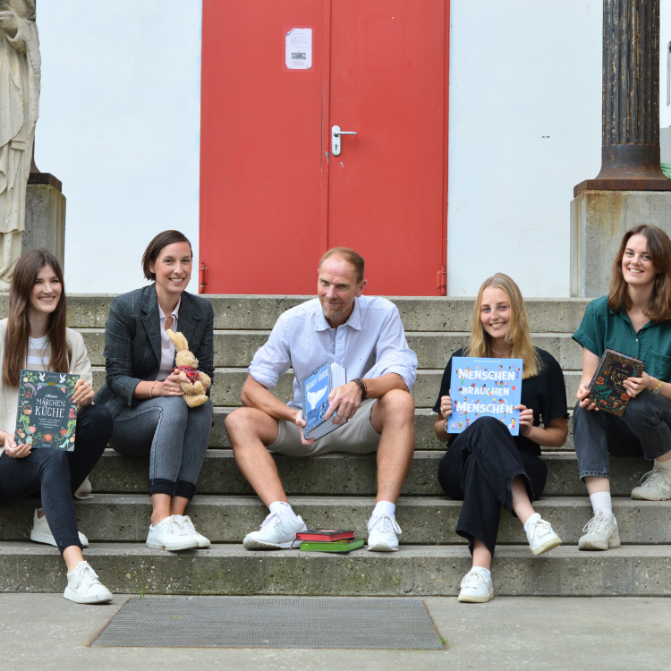 Ein Team aus jungen Menschen sitzen auf einer Treppe und halten Bücher hoch