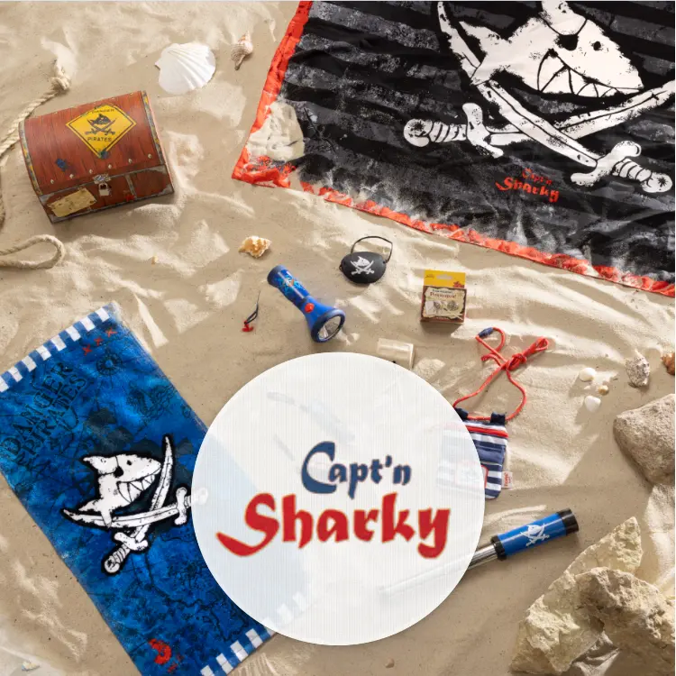 Auf einem Sandboden liegen verschiedene Produkte von Capt'n Sharky, unter anderem Ein Handtuch, eine Taschenlampe und ein Fernrohr