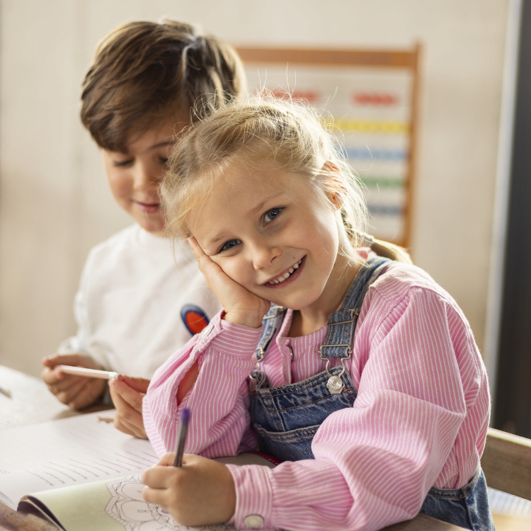 Mädchen sitzt an Schreibtisch mit einem Stift in der Hand und lächelt, ein Junge sitzt ebenfalls mit Stift hinter ihr