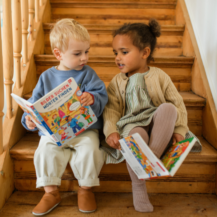 Zwei Kinder sitzen auf einer Holztreppe und schauen sich Pappbücher an